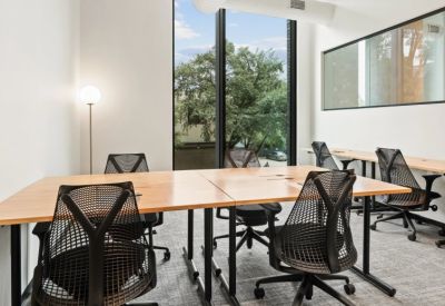 Open-plan workspace with light wood desks and ergonomic black mesh chairs by a large window.