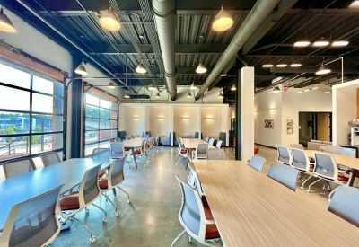 Spacious communal coworking area with industrial ceilings, long tables, and large glass garage doors.