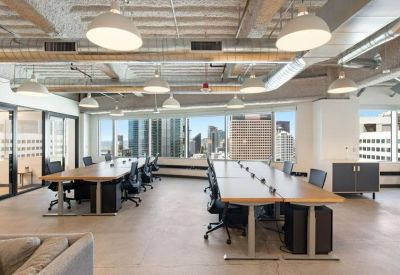 Bright open-plan office with long wooden desks and large windows overlooking the city skyline.