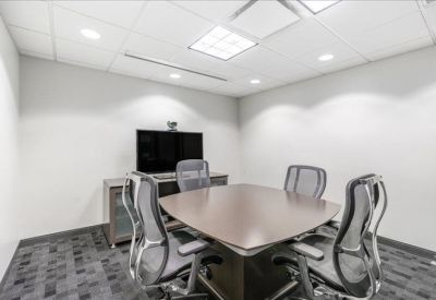 Small meeting room with a square wood-topped table, four grey mesh chairs, and a wall-mounted monitor.