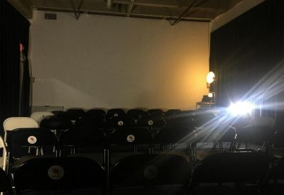 Dark screening room with rows of black chairs facing a large white projector screen.
