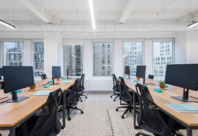 Bright open-plan workspace with multiple wooden desks, black mesh chairs, and large windows.