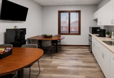 Bright communal kitchen and breakroom featuring white cabinetry, dining tables, and a wall-mounted TV.