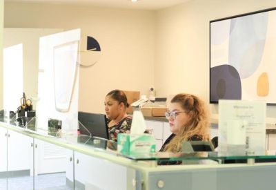 Two receptionists working at a modern white and glass front desk in a bright lobby area.
