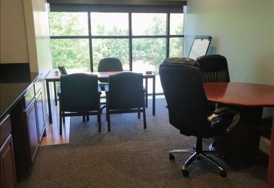 Private office with a wood-top desk, ergonomic chairs, and a large window overlooking trees.