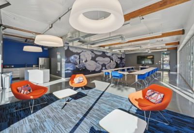 Spacious breakout area with orange chairs, blue rug, and a kitchen area.