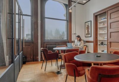 Lounge area with velvet chairs and a woman working at a round table near large windows.