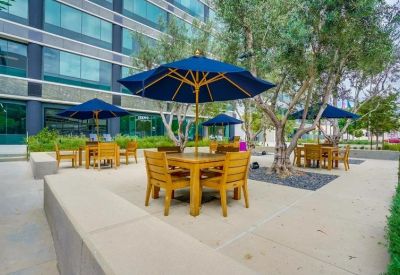 Outdoor patio area with wooden tables, blue umbrellas, and lush green trees.