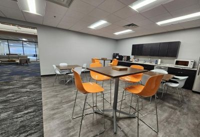 Communal breakroom with high-top wooden tables, bright orange chairs, and a kitchenette area.