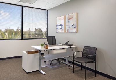 Bright corner office with a sit-stand desk, guest chair, and large windows with blinds.