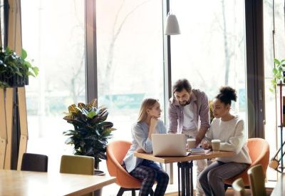 A small group collaborates around a laptop at a bright table near large windows.