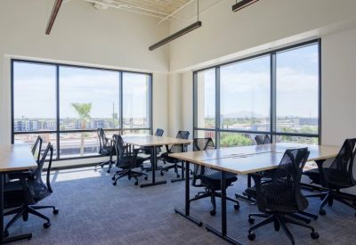 Sunlit open-plan office with several light-wood desks and black mesh chairs facing large windows.