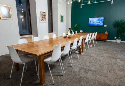 Long wooden boardroom table with white chairs set against a dark green feature wall with a TV.