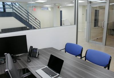 Modern private office suite with a dark wood desk, laptop, and blue chairs near a glass wall.
