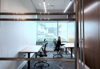 View through a glass partition of a shared office with workstations and a marble-patterned floor.