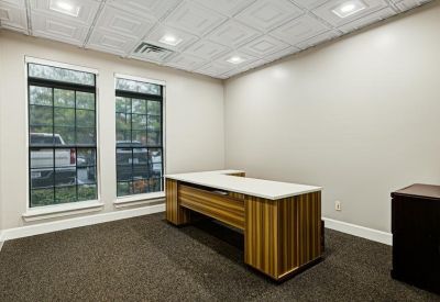 Private office with L-shaped wood-grain desk and large windows overlooking trees.