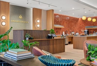 Reception area with a wood-slat desk, Industrious logo on a gray wall, and lush indoor plants.