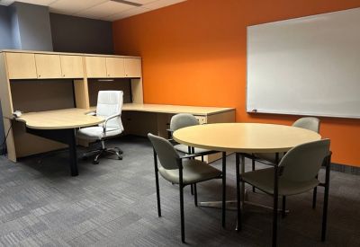 Meeting room with a round table, four grey chairs, and a large whiteboard on an orange wall.