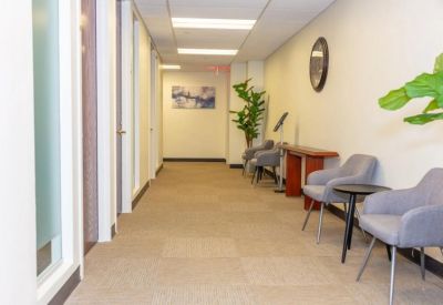 Well-lit office corridor with grey armchairs, small side tables, and potted plants for a welcoming feel.