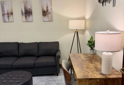 Office lounge area with a dark sofa, tripod floor lamp, and a view of a wooden desk and chair.