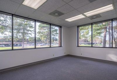 Spacious corner office with grey carpeting and large windows overlooking trees.