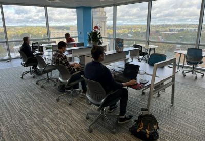 Open-plan office with employees working at long desks facing expansive windows with city views.