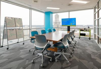 Meeting room with a wooden table, blue chairs, whiteboard panels, and large corner windows.