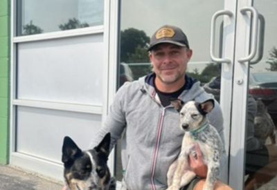 A man smiling with two dogs in front of the building's glass entrance.
