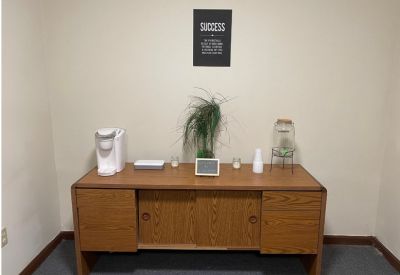 Communal coffee and water station on a wood-grain sideboard with a motivational poster.