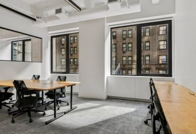 Bright shared office space with wooden desks, black mesh chairs, and large windows.