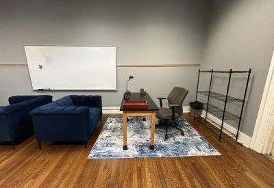 Office space with a blue velvet sofa, wooden desk on a blue abstract rug, and a tall wire shelf.