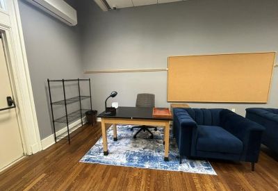 Office area showing a desk with a task lamp, a blue armchair, and a large cork board on a grey wall.