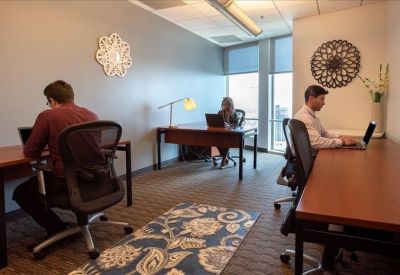 Brightly lit shared office space with wooden desks, ergonomic chairs, and a blue patterned rug.