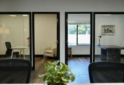 Private office suites with glass doors seen from behind a workstation area with greenery.