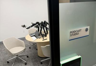 Professional podcast studio featuring a round wooden table with multiple microphone arms and a digital booking screen.