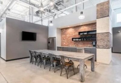 Communal breakroom with a long wooden harvest table, brick feature wall, and kitchen area.