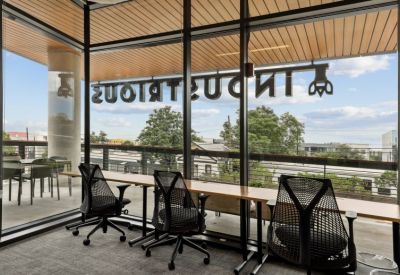 Modern workspace with wooden desks and ergonomic chairs overlooking a leafy balcony through large windows.