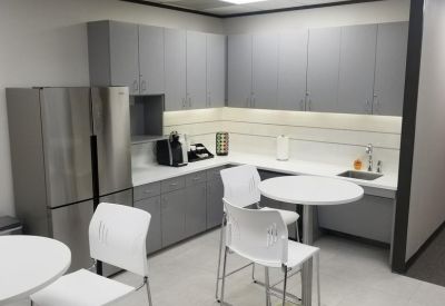 Modern communal kitchen and break area with grey cabinetry and white bistro seating.