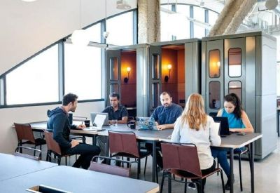 Open-plan workspace with a long communal table, privacy phone booths, and people working on laptops.
