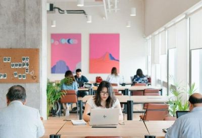 Open-plan office with people working at long wooden desks under bright lighting and pink wall art.
