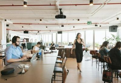 Open-plan office area with long communal desks, ergonomic chairs, and industrial ceiling details.