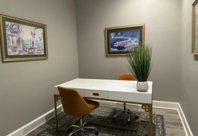 Bright two-person office with a white desk, orange chairs, and framed artwork.
