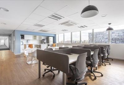 Open-plan workspace with rows of desks, ergonomic chairs, and a kitchenette in the background.