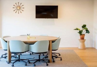 Minimalist meeting room featuring a round wooden table, light blue chairs, and a wall-mounted screen.