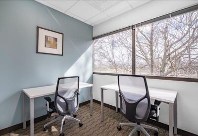 Bright corner private office with two desks and large windows overlooking trees.