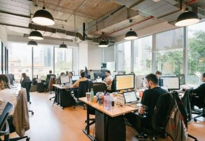 Sunlit open-plan office with rows of wooden desks, ergonomic chairs, and industrial ceiling details.