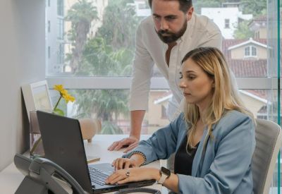 A man and woman collaborating over a laptop in a bright office with green hillside views.