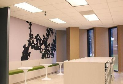 Bright communal break area with a long white table, green cushioned bench, and tree silhouette mural.