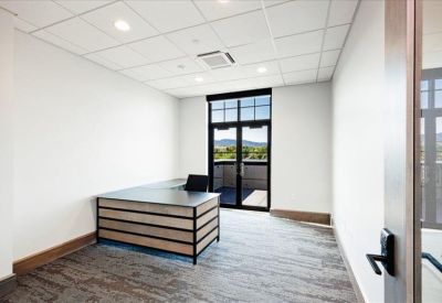 Private office with a light wood desk, grey carpet, and a glass door leading to a balcony.