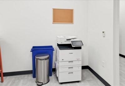 Communal print station with a white copier, blue recycling bin, and a wall-mounted corkboard.